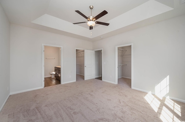 unfurnished bedroom with a walk in closet, ensuite bath, a raised ceiling, carpet floors, and ceiling fan