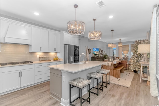 kitchen with white cabinets, light countertops, an island with sink, decorative light fixtures, and custom range hood