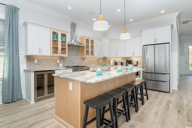 kitchen featuring beverage cooler, wall chimney range hood, a center island with sink, white cabinets, and stainless steel refrigerator