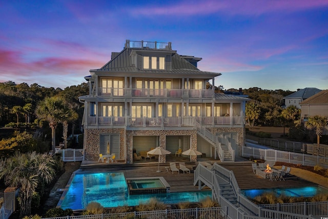 back house at dusk with a swimming pool with hot tub, a balcony, an outdoor fire pit, and a patio area