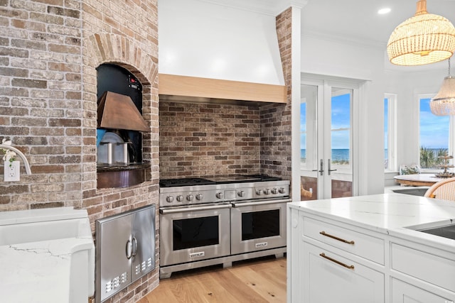 kitchen featuring premium range hood, range with two ovens, hanging light fixtures, light stone countertops, and white cabinetry