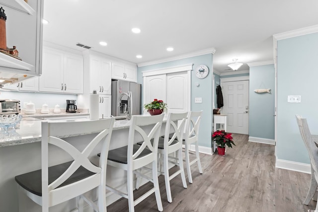 kitchen with white cabinets, stainless steel refrigerator with ice dispenser, a kitchen bar, ornamental molding, and light stone counters