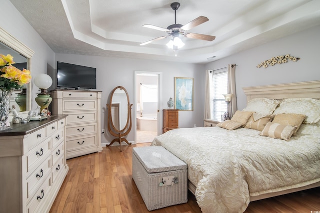 bedroom featuring light wood-type flooring, a raised ceiling, ensuite bath, and ceiling fan