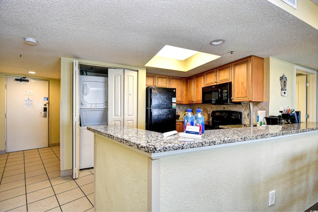 kitchen with light stone counters, kitchen peninsula, stacked washer / drying machine, light tile patterned flooring, and black appliances