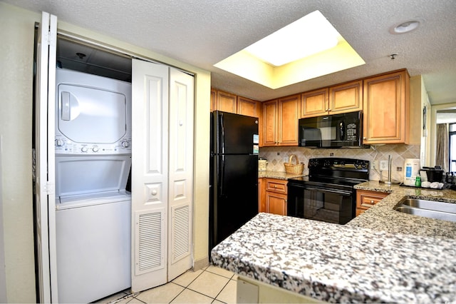 kitchen featuring stacked washer and clothes dryer, black appliances, tasteful backsplash, light tile patterned flooring, and kitchen peninsula