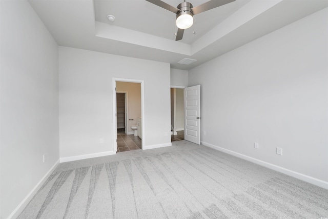 unfurnished bedroom with ceiling fan, a raised ceiling, light colored carpet, and connected bathroom