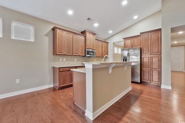 kitchen with a kitchen island with sink, a kitchen breakfast bar, decorative backsplash, appliances with stainless steel finishes, and light stone counters