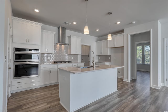 kitchen featuring wall chimney exhaust hood, sink, pendant lighting, white cabinetry, and an island with sink