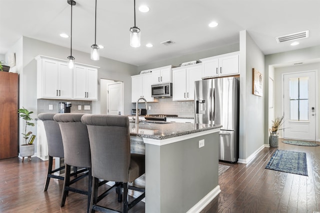 kitchen featuring a kitchen bar, stainless steel appliances, dark stone countertops, white cabinetry, and an island with sink