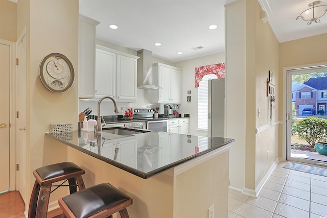 kitchen with kitchen peninsula, a kitchen bar, wall chimney exhaust hood, stainless steel appliances, and white cabinetry