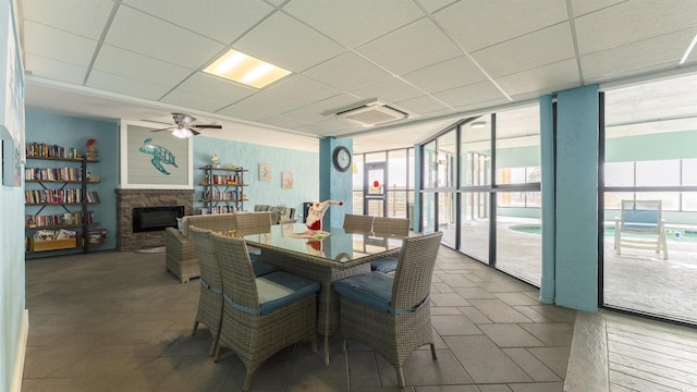 dining space featuring a drop ceiling, a stone fireplace, expansive windows, and ceiling fan