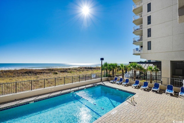 community pool with a patio area, a beach view, a water view, and fence