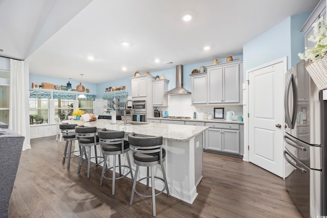 kitchen with hanging light fixtures, stainless steel appliances, wall chimney range hood, a kitchen bar, and a center island with sink