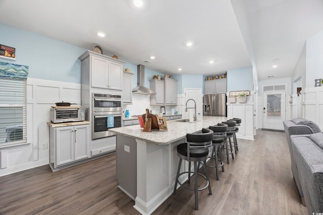 kitchen featuring wall chimney range hood, an island with sink, appliances with stainless steel finishes, light stone counters, and a breakfast bar area