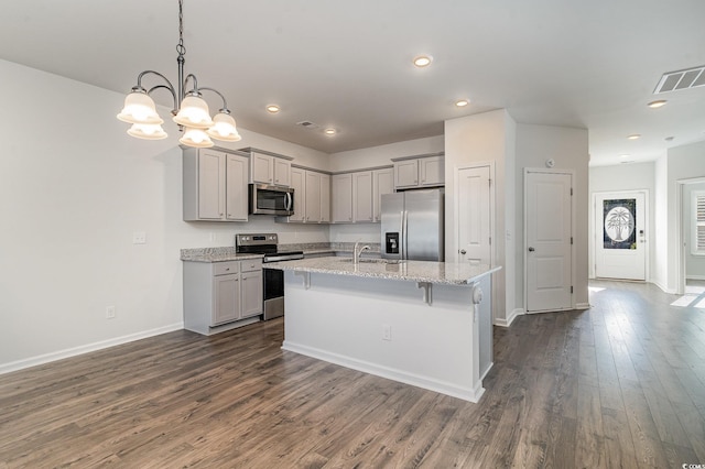 kitchen featuring hanging light fixtures, stainless steel appliances, light stone counters, an island with sink, and gray cabinets