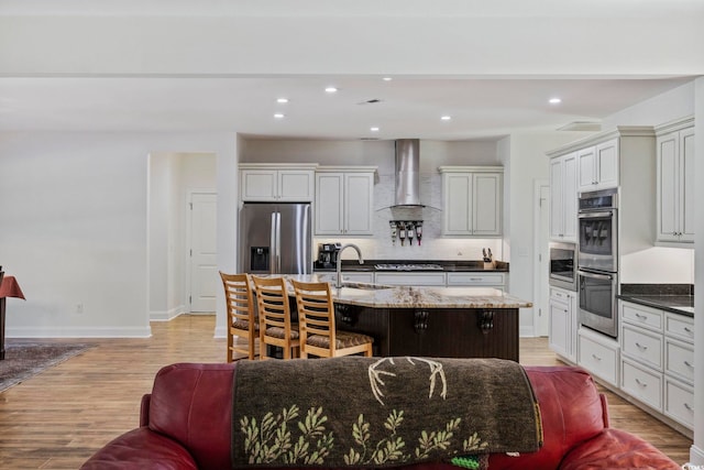 kitchen with wall chimney exhaust hood, appliances with stainless steel finishes, an island with sink, and a sink