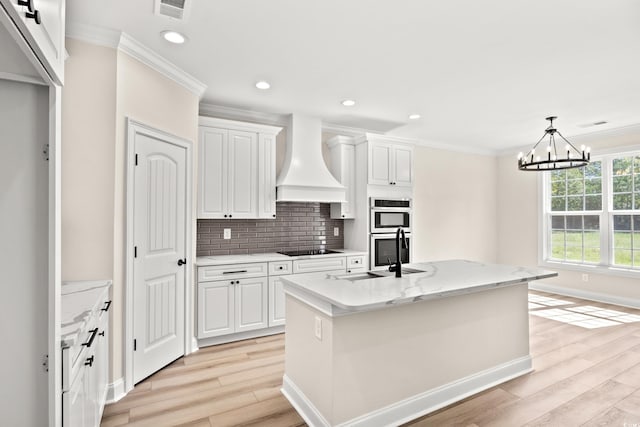 kitchen with stainless steel double oven, white cabinetry, premium range hood, and a kitchen island with sink