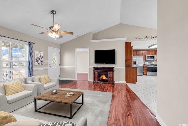 living area featuring baseboards, light wood-style flooring, ceiling fan, a lit fireplace, and vaulted ceiling