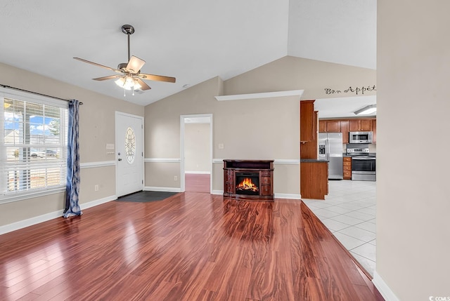 living room featuring a warm lit fireplace, baseboards, ceiling fan, wood finished floors, and vaulted ceiling