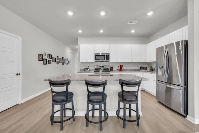 kitchen with a center island with sink, appliances with stainless steel finishes, light stone countertops, white cabinets, and light hardwood / wood-style flooring