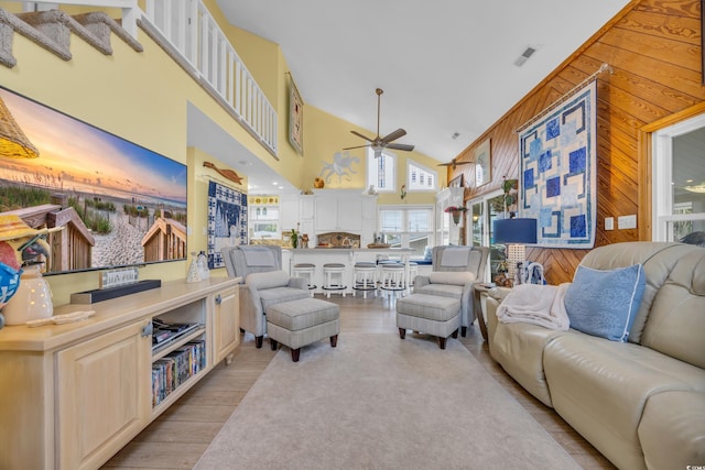 living room with ceiling fan, wood walls, a high ceiling, and light hardwood / wood-style flooring