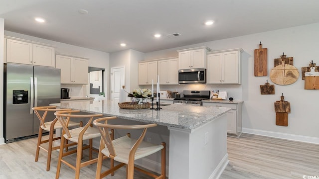 kitchen with stainless steel appliances, light wood-style flooring, decorative backsplash, an island with sink, and light stone counters