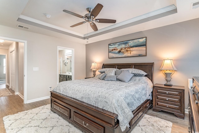 bedroom with crown molding, ceiling fan, ensuite bathroom, a tray ceiling, and wood-type flooring