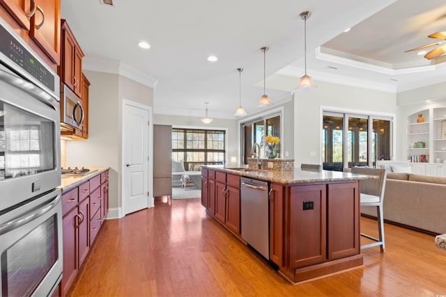 kitchen featuring a center island with sink, appliances with stainless steel finishes, decorative light fixtures, ornamental molding, and light stone counters