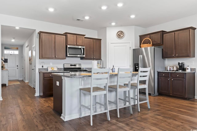 kitchen featuring dark hardwood / wood-style floors, stone counters, appliances with stainless steel finishes, a kitchen breakfast bar, and an island with sink