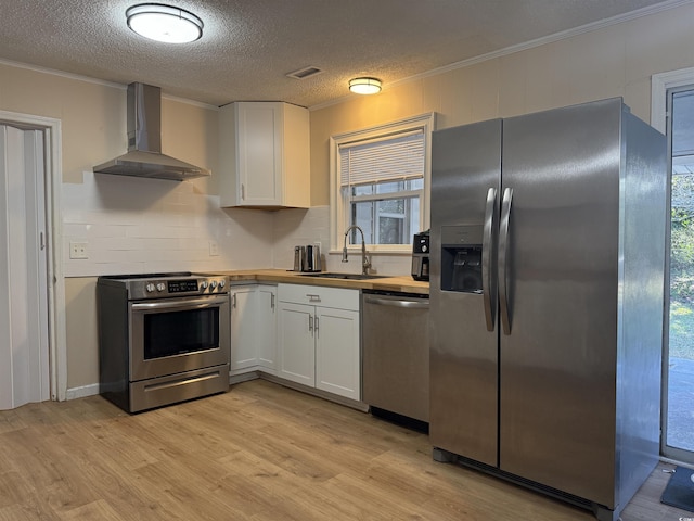 kitchen featuring ornamental molding, wall chimney exhaust hood, white cabinets, a sink, and appliances with stainless steel finishes