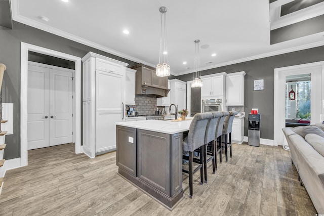 kitchen featuring a breakfast bar area, stainless steel oven, a center island with sink, hanging light fixtures, and white cabinets