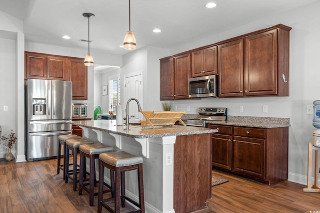 kitchen with pendant lighting, an island with sink, light stone counters, stainless steel appliances, and dark wood-type flooring