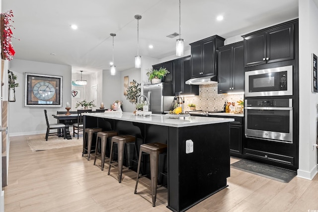 kitchen featuring a kitchen breakfast bar, light hardwood / wood-style flooring, pendant lighting, a kitchen island with sink, and stainless steel appliances