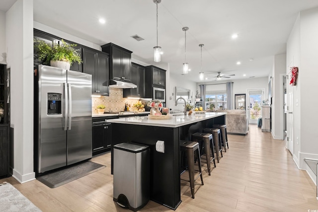 kitchen featuring light hardwood / wood-style floors, appliances with stainless steel finishes, decorative light fixtures, an island with sink, and a breakfast bar area