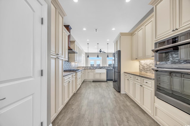 kitchen with pendant lighting, appliances with stainless steel finishes, cream cabinetry, dark stone counters, and light wood-type flooring