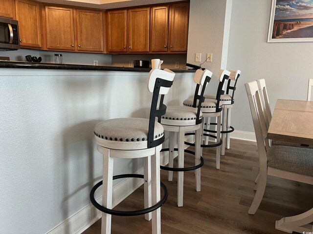 kitchen with dark countertops, stainless steel microwave, a breakfast bar, brown cabinets, and dark wood-style flooring