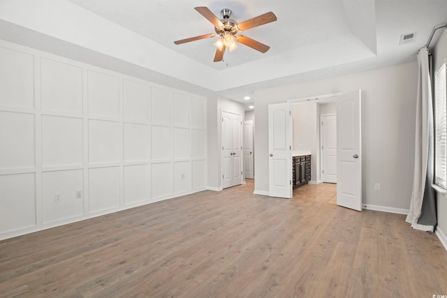 unfurnished bedroom featuring ceiling fan, connected bathroom, a tray ceiling, and light hardwood / wood-style floors