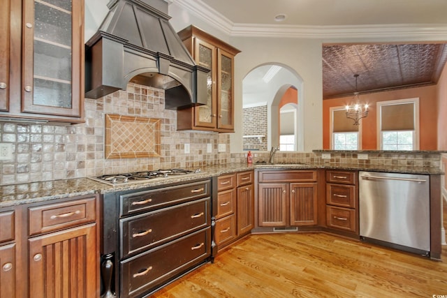 kitchen featuring brown cabinetry, ornamental molding, custom range hood, appliances with stainless steel finishes, and light wood-type flooring