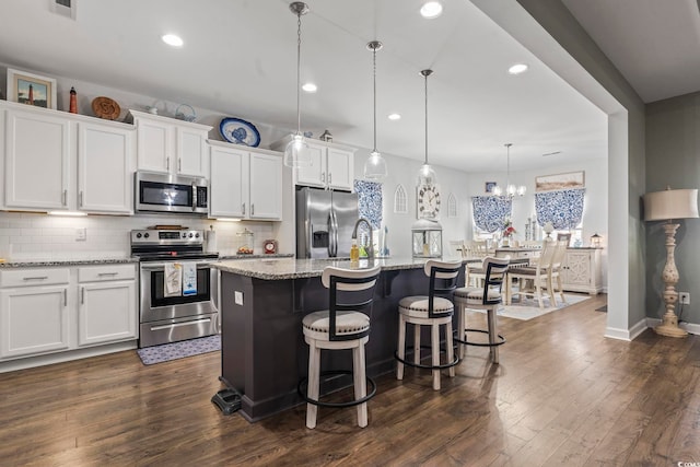 kitchen featuring appliances with stainless steel finishes, hanging light fixtures, light stone counters, white cabinets, and a center island with sink