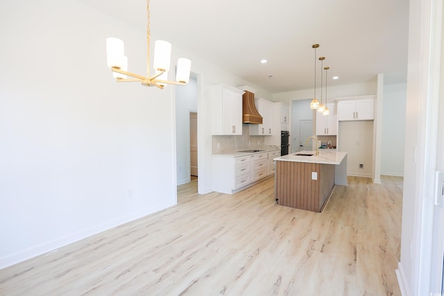 kitchen with sink, hanging light fixtures, custom range hood, white cabinets, and a center island with sink