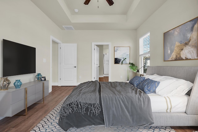 bedroom with a raised ceiling, visible vents, ceiling fan, ensuite bath, and wood finished floors