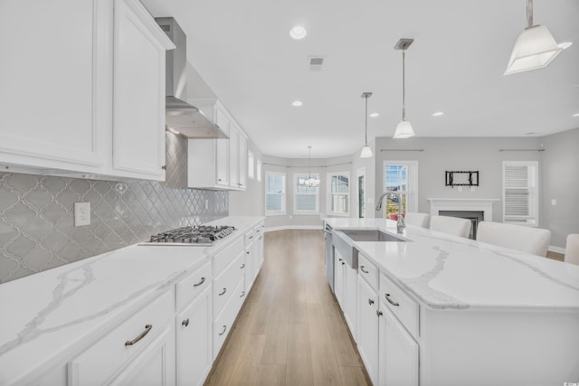 kitchen featuring decorative light fixtures, white cabinets, a large island with sink, stainless steel appliances, and wall chimney exhaust hood