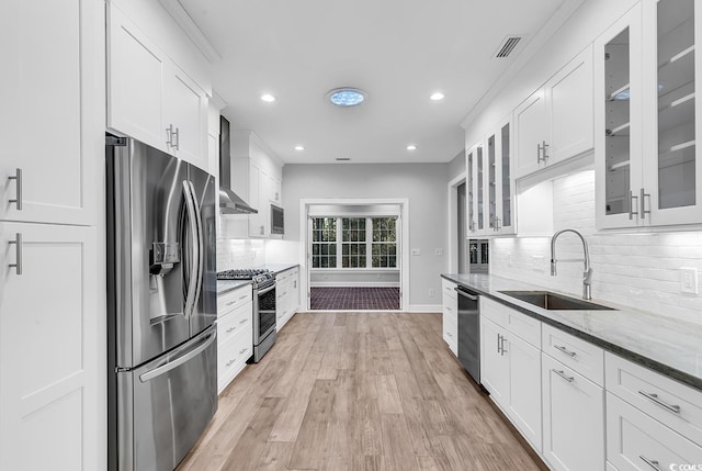 kitchen featuring appliances with stainless steel finishes, glass insert cabinets, white cabinetry, a sink, and wall chimney exhaust hood