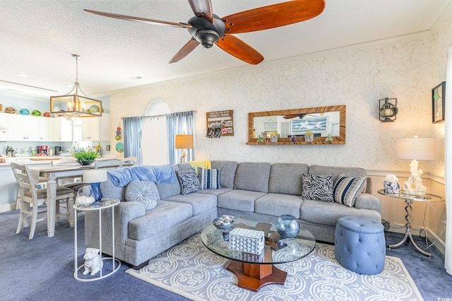 living room featuring wallpapered walls, ornamental molding, dark carpet, and ceiling fan with notable chandelier