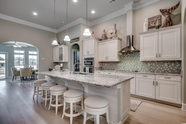 kitchen featuring white cabinetry, wall chimney exhaust hood, decorative light fixtures, and an island with sink