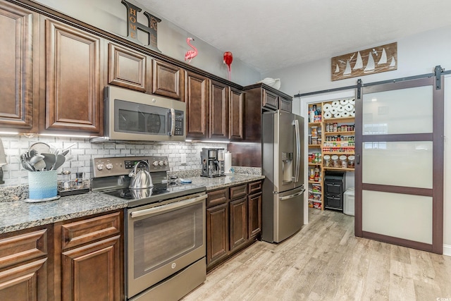 kitchen with tasteful backsplash, light wood-type flooring, stainless steel appliances, a barn door, and light stone countertops