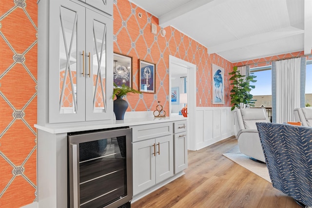 bar featuring wine cooler, beam ceiling, a wainscoted wall, light wood finished floors, and wallpapered walls