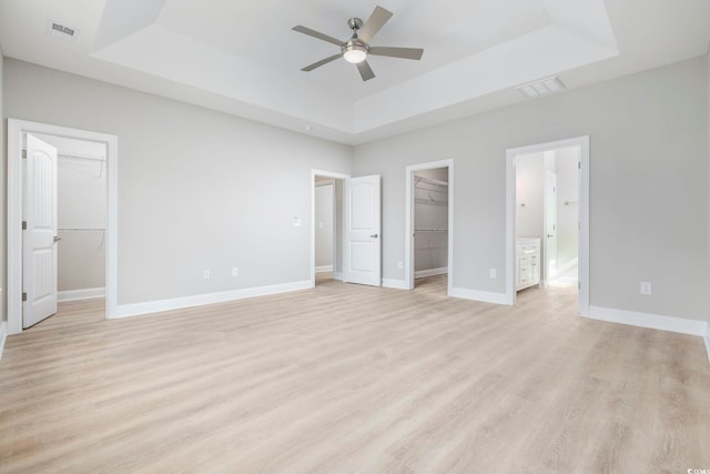 unfurnished bedroom with a walk in closet, a tray ceiling, a closet, ceiling fan, and light hardwood / wood-style floors