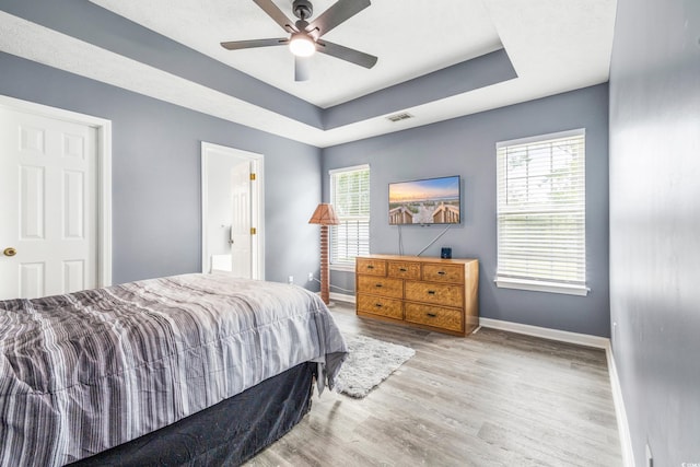 bedroom with ensuite bath, light hardwood / wood-style flooring, a raised ceiling, and ceiling fan