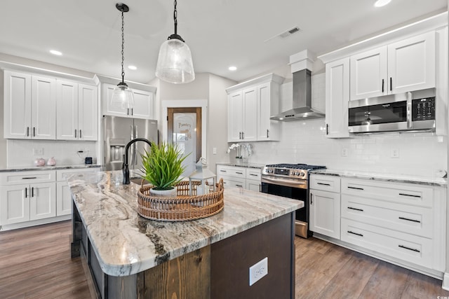 kitchen featuring a kitchen island with sink, stainless steel appliances, white cabinetry, hanging light fixtures, and wall chimney exhaust hood